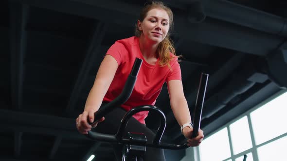 Athletic Girl Performing Jumping Aerobic Riding Training Exercises on Cycling Stationary Bike in Gym