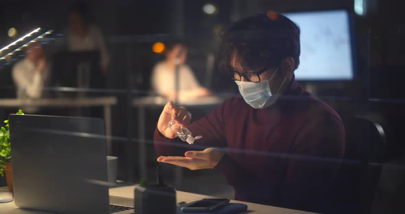 Asian Businessman Working in Office at Night Wearing Face Mask Sitting at Desk Using Hand Sanitizer