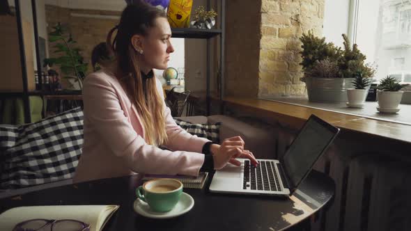 Beautiful Young Caucasian Woman Drinking Coffee and Typing on a Keyboard Inside a Cafe at a Wooden