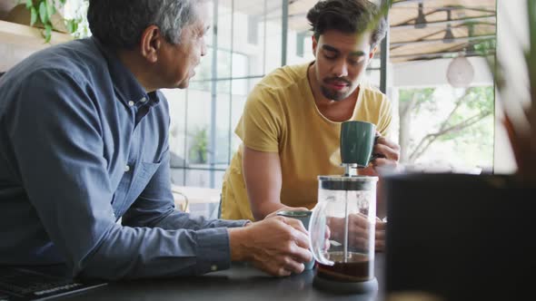 Man talking to young son drinking coffee from mug while leaning on kitchen counter at home