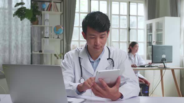 Young Asian Man Doctor Is Using Tablet While Work With Laptop Computer In Workplace