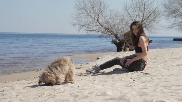 Brunette in Top and Jeans Sits on Sand Sea Beach with Dog