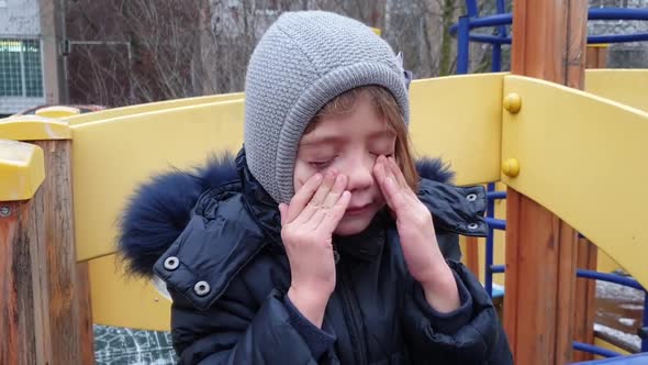 close up view of little girl rubbing her eyes with her hands and crying on playground in the fall