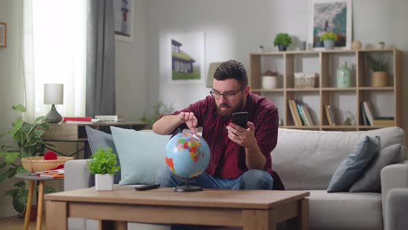 Young Handsome Guy Examines the Globe with a Phone in His Hands While Sitting on the Sofa in the