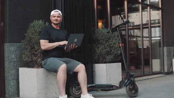 Adult man well dressed with electric scooter working with his laptop in the street in the city. A yo