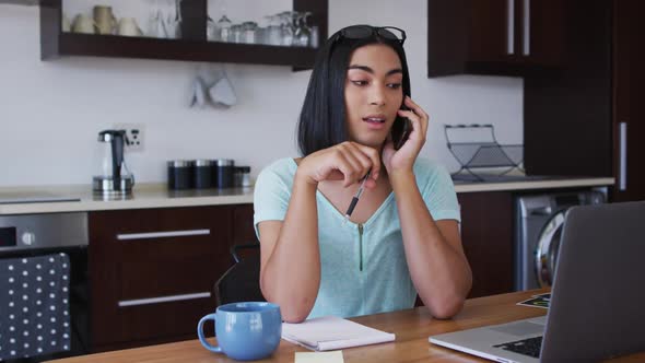 Mixed race gender fluid person sitting at desk working from home using a laptop and talking on smart