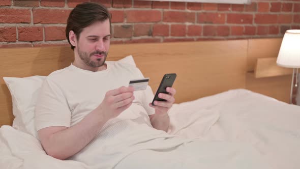 Casual Young Man Making Online Payment on Smartphone in Bed