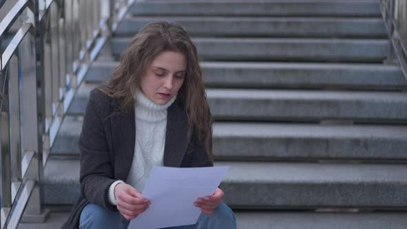 Young Brunette Caucasian Sad Woman Reading Letter Tearing Paper Sitting on Urban City Stairs