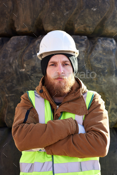 r wearing hardhat standing with arms crossed and looking at camera against huge truck tires