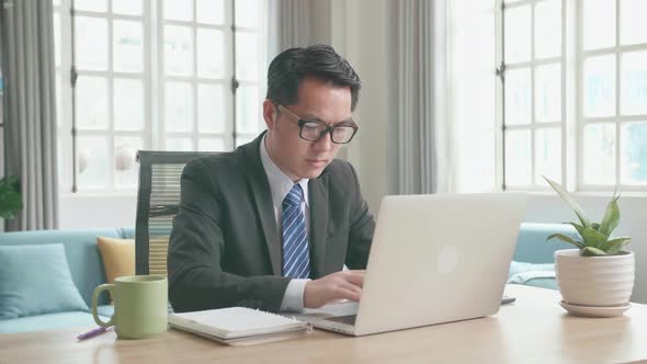 Asian Businessman With Glasses Wearing Business Suit Typing On Computer While Working At Home