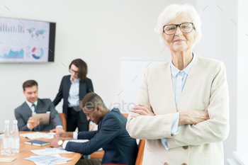 e officer in glasses wearing jacket standing in meeting room and looking at camera while business analysts working with data in background.