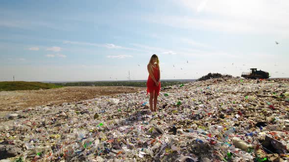 Epic Dolly in Young Woman in Red Dress Standing at Huge Trash Dump with Many Seagulls in Background