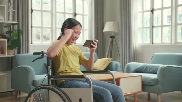 Excited Asian Kid Girl Sitting On Wheelchair Playing Video Game On Mobile Phone And Celebrating alt