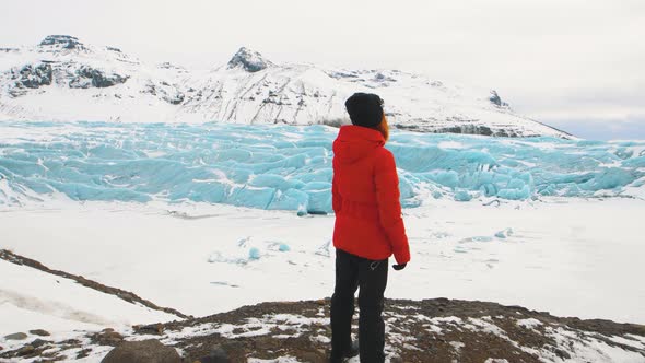 Young Woman Tourist in a Red Jacket Walks Around Huge Glacier in Iceland