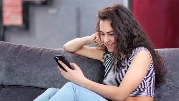 Beautiful Pleased Young Woman Enjoying Chatting Using Smartphone Relaxing at Home