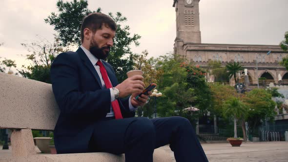 Mature Business Man Reading a Newspaper Sitting in the Park on the Bench