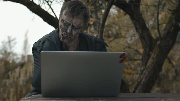 Portrait of Male Zombie Using Laptop Sitting at Wooden Table Outdoors