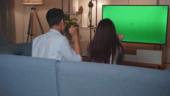 Back View Of Excited Asian Couple Watching Tv With Mock Up Green Screen Together Sitting On A Sofa