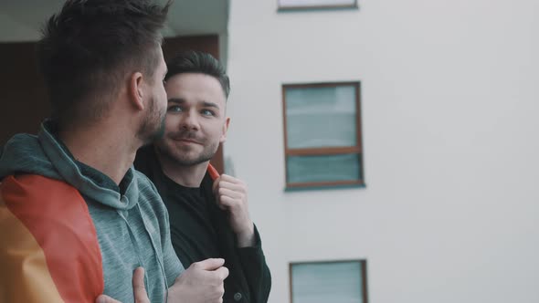 Proud Lovely Gay Male Couple Standing on the Balcony Covered with Pride Rainbow Flag