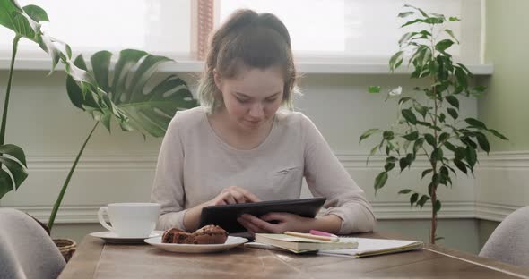 Teenager Girl Sitting at Home at Table Eating and Reading, Using Digital Tablet for Learning