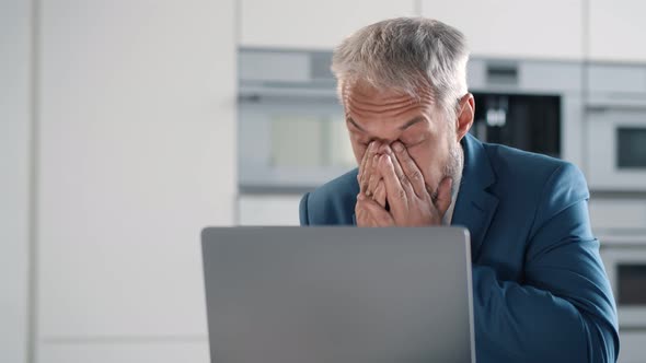 Exhausted Mature Businessman Working in Home Office with Laptop Sitting in Kitchen