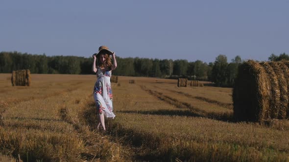 Beautiful Redhaired Woman in a White Dress and Hat Walks Away on the Background of the Field