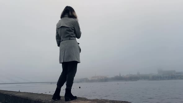 Girl standing near misty river and looking how city disappears, meanwhile vaping