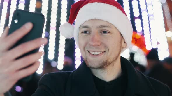 Cheerful Man Taking Selfie and Builds Funny Faces on Mobile Phone, Dressed in Christmas Hat