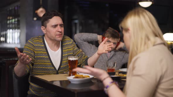 Angry Couple of Parents Arguing in Restaurant with Blurred Teenage Son in Headphones Sitting at