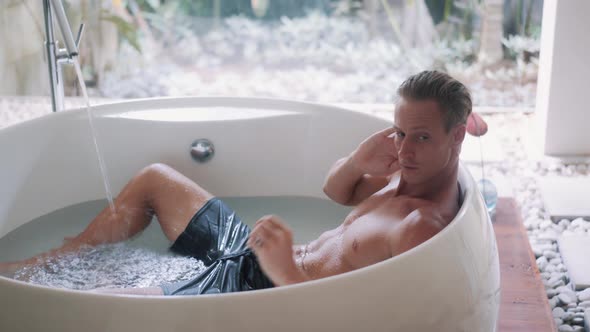 Young Sporty Guy with Muscular Body Rests in Large Bathtub at Home