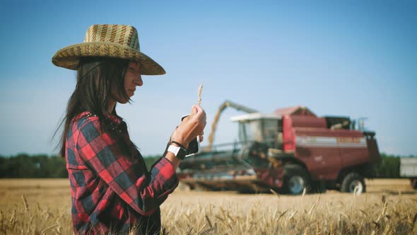 Young Woman Farmer in Hat Standing Against the Background of a Working Combine Harvester at Sunset