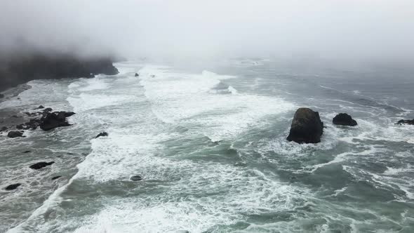 White foamy emerald green waves crash on a rocky jagged fog shrouded coastline, aerial