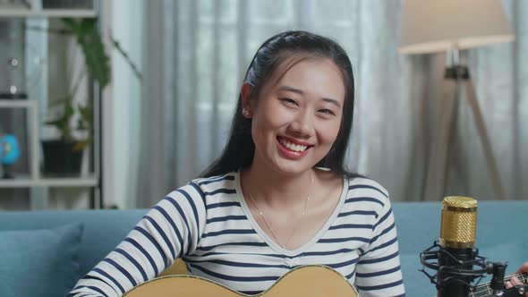 Close Up Of Asian Woman Playing A Guitar And Smiling To Camera While Live Stream At Home