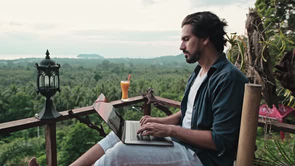 A young, handsome man with a beard works on a laptop in a cafe overlooking an incredible sunset.