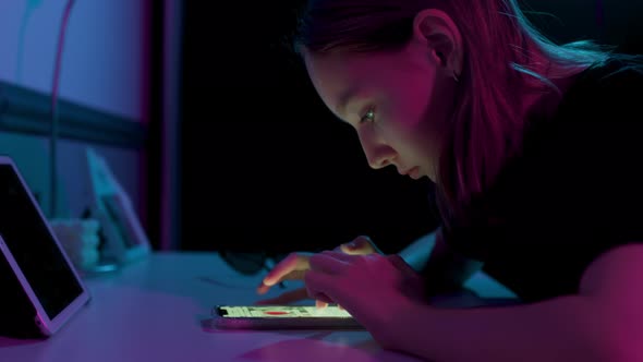 Teenage Girl Typing Text Message on Mobile Phone Sitting at Table at Night Indoors in Purple Pink