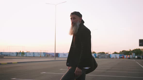 Stylish Handsome Middleaged Man with Long Gray Beard Standing with Longboard During Sunset on Urban