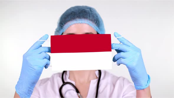 Close-up. Doctor in Glasses, Blue Medical Cap, Gloves Holds in Hands Medical Mask with Monaco Flag