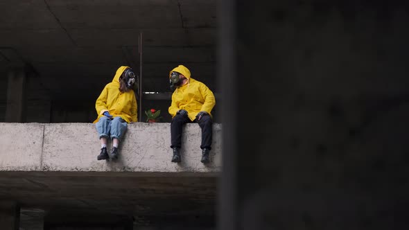 Man and Woman are Sitting on Edge of Abandoned Building in Gas Masks and Yellow Protective Suits in