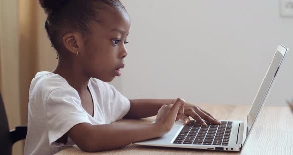 Afro American Preschool Girl Typing Keyboard of Laptop Computer Sitting at Table at Home. Child