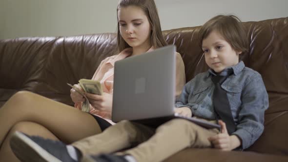 Portrait Older Sister and Younger Little Brother Sitting on the Leather Sofa. The Boy Holding Laptop