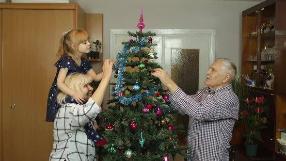 Kid Girl with Senior Grandmother and Grandfather Decorating Artificial Christmas Tree with Toys