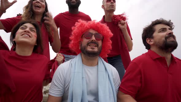 A Group of Soccer Team Fans Celebrate Victory in the Stands Wearing Red Jerseys While a Fan of the