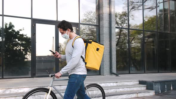 Courier with Yellow Backpack and Protective Mask Walking with Bicycle in City Center and Using