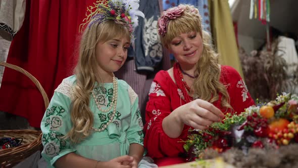 Smiling Ukrainian Mother and Curios Cute Daughter Sitting Indoors Making Traditional Head Wreath in