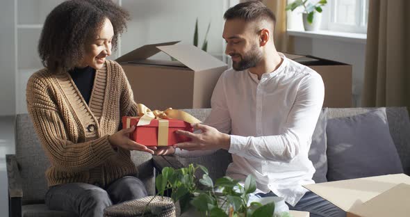 Excited Young African Girlfriend Receiving Gift From Caucasian Boyfriend at Home, Happy Millennial