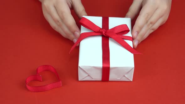 Girl Hands Put White Gift Box with Red Bow on Red Table Decorated with Heart