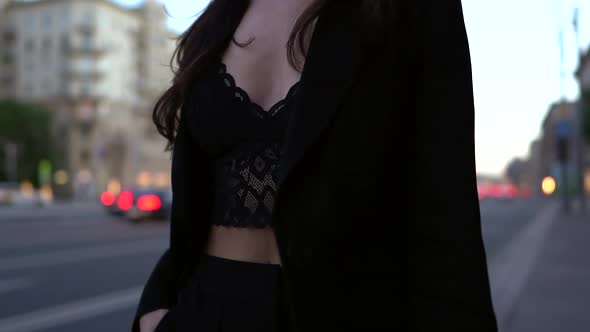 a Young Brunette in Dark Clothing Walks Along a City Street at Twilight Against the Background of