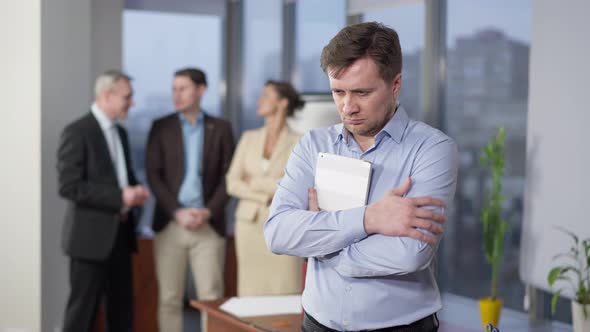 Portrait of Stressed Overworked Man Holding Tablet Looking Back at Blurred Colleagues Talking