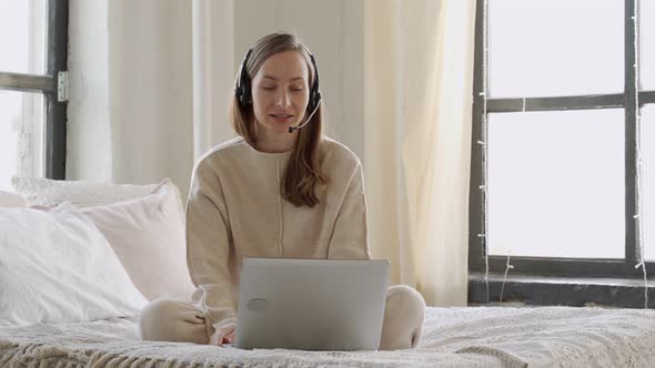 Smiling Woman Wearing Headset Looking at Laptop Sitting on Her Bed with Laptop Making Video Call