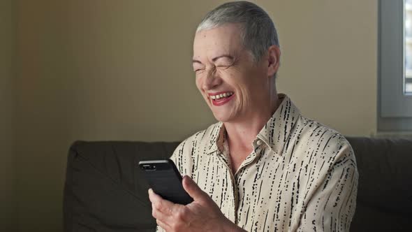 An Elderly Woman Looks at the Smartphone Screen and Laughs Very Cheerfully and Sincerely